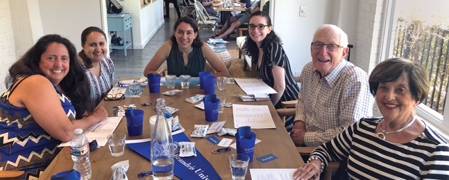 Five women and a man sit around a long table covered with blue Brandeis swag items.